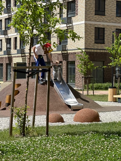 In Znamya Oktyabrya, Russia, a modern children's playground is depicted in front of a brick apartment building. A man in a white t-shirt and blue pants is standing near a slide, holding a red and brown object. A young child in a striped shirt and pants is on the slide. The playground features a brown-surfaced mound with wooden steps and a metal slide. The ground is covered with white gravel and green grass with small white flowers. Trees with green leaves are present, and a wooden trash can and bench are visible. The apartment building is made of beige and brown bricks with dark-framed windows.