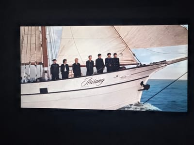 The image displays a white-hulled sailboat named "Arirang" underway on blue water under a partially cloudy sky. Six young adult males, dressed in dark, uniform-style attire, stand in a line on the starboard side of the boat's foredeck, facing generally forward. The boat exhibits a prominent mast and rigging with furled or partially furled sails, and a visible anchor on its bow. A wake is present at the boat's bow, indicating forward motion through the water. This scene is situated in Casa Blanca, Mexico.