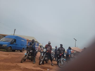 The image depicts an outdoor scene in what appears to be a semi-urban or rural area in Tangu, Nigeria. A group of men are gathered around several motorcycles on a dirt road. The road surface is reddish-brown and appears to be uneven and somewhat muddy in places.

In the foreground, a blue van is parked. It has "GARI YAWAYE" and "PALANG OIL LTD." written on its side. Behind the van and to the right, several men are standing or sitting on motorcycles.

The first man on the left, in the foreground, is wearing a blue jacket with black stripes and a checkered scarf around his neck. He is leaning against a motorcycle. He has dark skin and short, dark hair, and is wearing green boots.

To his right, another man, wearing a maroon long-sleeved shirt with "US" printed on it, is standing next to his motorcycle. He is wearing dark pants and flip-flops. He has a light-colored knitted cap on his head.

Further to the right, a man in a brown jacket and a dark cap is sitting on a red and blue motorcycle. He has dark skin.

Another man in a blue jacket is standing near the center-right of the image, next to a motorcycle.

The background shows some buildings, including one with a light-colored roof and signs that are difficult to read. There are also utility poles and overhead wires visible. The sky is overcast and a pale gray. The overall atmosphere suggests a moment of rest or waiting.