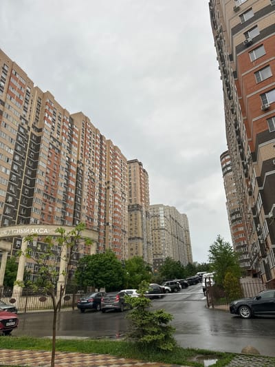 The image depicts a street scene in Kuleshovka, Russia, showcasing tall apartment buildings on either side. The sky is overcast. On the right side of the frame, cars are parked along the curb, interspersed with trees and greenery. A sidewalk with a patterned surface borders the street. In the foreground, there is a fence and a sign with Cyrillic text ("КРАСНЫЙ АКСАЙ"). The architectural style of the buildings suggests a modern urban environment.
