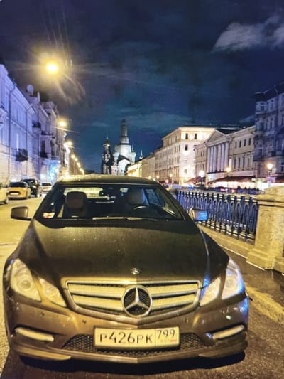 This media file shows a Mercedes-Benz car parked on a city street at night. The car is dark-colored and faces the camera, with its headlights illuminated. The license plate is visible and reads "P426PK 799 RUS".

The scene depicts a street with buildings lining both sides, illuminated by streetlights and the lights from the buildings. The architecture suggests a European city, likely in Russia given the license plate. There are other cars and a fence visible in the background, as well as a domed church spire in the distance. The sky is dark with some clouds and a bright street lamp in the upper left corner casts light rays. The road appears wet, suggesting recent rain.

The time of day is clearly night, indicated by the dark sky and artificial lighting. No people are prominently featured in the image. The overall mood is urban and nocturnal. The presence of the car and the streetscape implies a public space, possibly a road or a promenade.