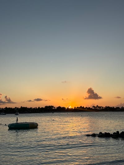 The image depicts a sunset over the water in Himmafushi, Maldives. The horizon features a dark silhouette of a landmass, densely lined with palm trees. The sun, partially obscured by these trees, is setting, casting a bright orange-yellow glow and emitting visible sun rays across the water. The sky transitions from a clear, light blue at the top to vibrant orange and yellow hues near the horizon, with several scattered clouds, some reflecting the sunset colors. A faint crescent moon is visible in the upper-left quadrant of the sky.
In the mid-ground, a large, round, inflatable water trampoline, predominantly green with a yellow edge, floats on the calm, reflective water. A single individual stands centrally on the trampoline's surface, silhouetted against the bright horizon. Two additional human figures, also dark silhouettes, are partially submerged in the water to the left of the inflatable structure. Several dark buoys are visible in the water. To the far right in the foreground, a cluster of dark rocks protrudes from the water. Multiple small boats are moored in the distance, near the left side of the horizon line.