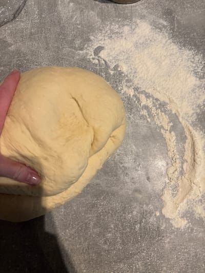 A close-up, top-down view captures a human hand actively pressing into a substantial mass of pale yellow dough on a dark gray, speckled countertop, located in Groß-Gerau, Germany. The dough, exhibiting a smooth yet pliable texture with visible folds from manipulation, occupies the lower-left and central portions of the frame. The left hand, with fair skin and an index finger displaying a clear or light-colored manicured nail, applies pressure to the dough, indicating a kneading or shaping action. The dark gray work surface is dusted unevenly with white flour, particularly concentrated in an arcing pattern across the upper-right and central-right areas, providing a non-stick medium for the dough. A faint, out-of-focus translucent object, possibly plastic wrap or a container edge, is barely perceptible in the upper-left corner.
