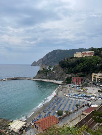 The image depicts a high-angle view of the coastal town of Monterosso al Mare, Italy, under an overcast sky. The Ligurian Sea occupies the left and central-left, with a curved concrete breakwater extending from a rocky promontory, forming a partially enclosed harbor. A red and white motorboat is visible in the water within this harbor.
The central area features Fegina Beach, a pebble and gravel shore. Hundreds of blue-striped sun loungers are arranged in orderly rows across the beach, accompanied by some matching umbrellas. Approximately 50 to 70 individuals are present on the beach; some are sunbathing on loungers, others walk along the shoreline, and a few are wading in the shallow water. Several red and white rescue buoys are visible on the sand.
On the rocky promontory to the left, a stone tower (Torre Aurora) stands, with a modern glass-fronted structure located below it. To the right of the beach, a paved dry dock area contains numerous small boats, most covered with blue tarpaulins. Behind this area, multiple multi-story buildings with red, yellow, and cream facades rise up the hillside, connected by visible stairs and paths. Further up the mountain, a large historic building complex with a red-tiled roof and pale walls (Convento dei Cappuccini) overlooks the scene. A smaller, dark stone tower is visible on a more distant peak.
Below the dry dock, a set of railway tracks runs parallel to the coast. In the immediate foreground, a building with a reddish-brown tiled roof is partially visible, featuring an outdoor dining area with a striped canopy. A white utility van is parked near the dry-docked boats. The environment indicates a coastal tourist destination with both natural features and developed infrastructure.