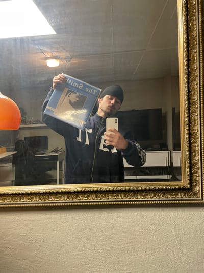 A male individual is depicted in a mirror selfie taken indoors in Oberhausen, Germany. He is positioned centrally, facing the camera reflected in an ornate gold-framed mirror. The individual wears a dark long-sleeved zipped hoodie with light-colored detailing on the front and sleeves, along with a black beanie. He holds a white smartphone with his right hand, angled to capture the reflection, and with his left hand, he holds a light blue vinyl record album above his head. The album cover features a monochrome image of a person and the reversed text "The Smiths." The reflection behind the individual shows an interior room with a textured light-colored wall below the mirror. Within the reflection, an overhead fluorescent light fixture is visible on a concrete-colored ceiling. To the left, an orange spherical lamp is partially visible. Further back in the reflection, there is a desk or table with a computer monitor, a dark chair, and a large dark-screen television above a white cabinet unit.