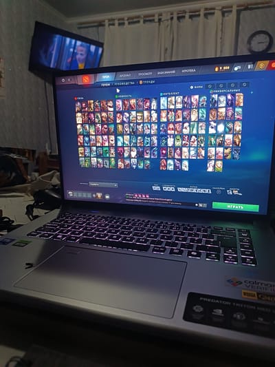 The image shows a close-up of a laptop screen displaying the game Dota 2, specifically the hero selection screen. The laptop is open and placed on a surface, likely a desk or bed, in a dimly lit room. The laptop's keyboard is illuminated with RGB lighting. In the background, a television screen is visible, displaying a distorted image of a person. The overall environment appears to be a residential setting, possibly a bedroom or living room. The image does not contain any specific visual cues that would allow for the identification of the city of Medveja, Moldova.