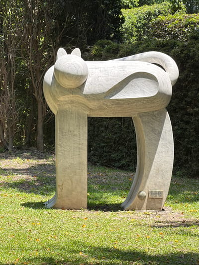 The image shows a large, abstract white marble sculpture in a park or garden setting, likely in Tehuixtla, Mexico. The sculpture consists of curved and rectangular elements arranged in a semi-circular form with a rounded protrusion on one side. It is positioned on a grassy area adjacent to dense green foliage and trees. The sculpture appears to be named "Mascacide," according to a small plaque. The lighting suggests it is either morning or late afternoon. There are no people visible.