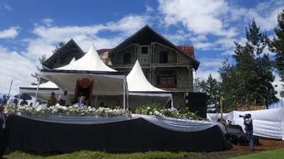 A large, white, tent-like structure, with a wooden chair in the center and a large, white cloth covering it. The structure appears to be situated in front of a large, unfinished, two-story brick building with a brown roof and large windows. There are trees in the background. Several people are sitting in front of the large, white, tent-like structure. A man in a dark shirt and black pants is standing in front of the chair, looking towards the camera, and a woman in a white dress is standing behind him. There are more people standing in the background, behind the tent. A man with a camera is standing to the right of the tent. The scene is likely a ceremony or event taking place in the city of Bukavu, Democratic Republic of the Congo, based on the provided coordinates.