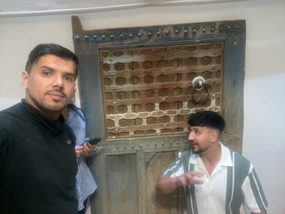 Two men pose with a dark wooden door featuring a central lattice panel and ornamental hardware. The man on the left wears a striped shirt, while the man on the right is in a black shirt. The scene is likely in Tassoultante, Morocco, based on the subject matter. The door displays aged details.