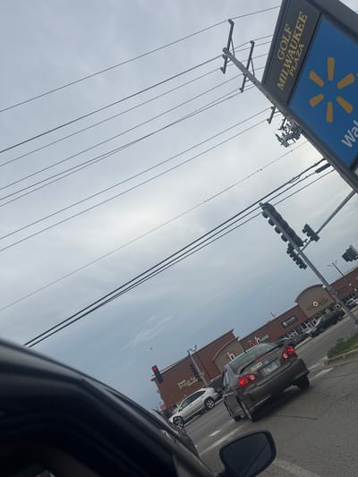 The image is taken from a vehicle at an intersection in Niles, United States of America, looking up and to the right. Overhead, a network of power lines crisscrosses the sky. To the right, a tall signpost displays "GOLF MILWAUKEE PLAZA" in gold lettering on a black background, next to a large blue sign with a yellow Walmart logo. In the distance, traffic lights are visible, one displaying a red light. Several cars are on the road, with a gray sedan in the foreground. Buildings are lined up along the street, including a storefront labeled "EGG N' HOLIC" and another with "Crumbl Cookies". The sky is overcast.