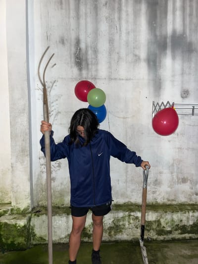 A person wearing a navy blue tracksuit and black shorts holds a pitchfork and shovel in what appears to be a garage or storage room. Several balloons are visible, one red balloon is attached to a metal extending structure on the wall, while the others are grouped on the wall. The walls are concrete with patches of mold and discoloration. The location is unknown, as the city of Florida, Argentina, is not identifiable from the visual information.