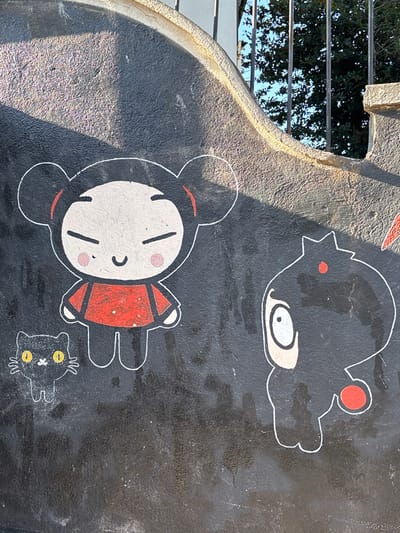 This image displays a section of a dark, textured wall featuring stylized cartoon graffiti art in Tomé, Chile. The primary figures include a character resembling Pucca on the left, depicted with a white face, a slight smile, rosy cheeks, and black hair in two buns with red accents, wearing a red tunic. Below this character, a small, black stylized cat with large yellow eyes and white whiskers is present. To the right, another character resembling Garu is shown in black, with a large white eye, a red dot on its head, and a red circular element on its lower right body. The wall exhibits a curved, lighter-colored coping along its upper edge, behind which dark vertical metal bars and green foliage are visible against a blue sky, indicating an outdoor setting. A distinct diagonal shadow from an external source falls across the upper left portion of the wall. The paint appears to have some minor wear and streaking on the dark background.