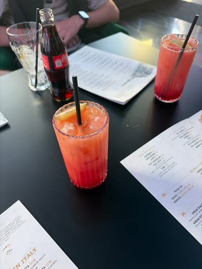 The scene is set on a black table, presumably at a restaurant, indicated by the presence of menus and drinks. Two cocktails in tall, fluted glasses with black straws are visible. One contains a red liquid and the other contains a pink liquid. Ice and a slice of lime are visible in the drinks. Beside one cocktail is a glass bottle of Coca-Cola. A partially visible arm with a watch suggests a person is seated at the table. A menu is unfolded, displaying the restaurant's offerings in the "TEAS & COFFEES" and "COLS BRINES" categories. The menu also includes alcoholic and non-alcoholic drink options such as Peroni and Heineken. Another menu section reads "IN ITALY" and lists pasta dishes. The text "HALAL" indicates dietary adherence to Islamic law. The location is in Leicester, United Kingdom, suggested by the menu content and the Coca-Cola bottle.