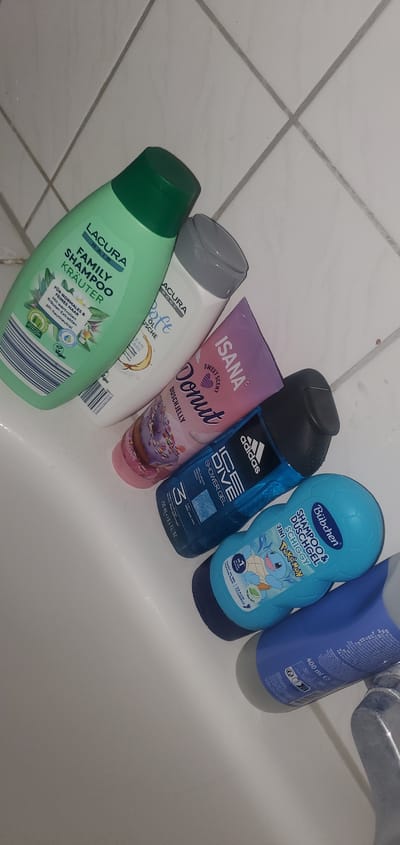 Six bottles of shampoo and shower gel are arranged in a bathtub. The bottles include Lacura Family Shampoo Kräuter, Lacura Body Soft Oil Dusche, Isana Sweet Scent Donut Duschjelly, Adidas Ice Dive Shower Gel, Bübchen Shampoo & Duschgel Schiggy (Pokémon themed), and another blue bottle of shampoo, possibly by Adidas, with "400 ml" visible. The background is a white tiled wall. The image was taken in Sasbach, Germany, in a bathtub.