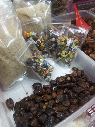 The scene, located in Lê Thanh Nghị Ward, Vietnam, depicts a retail food display. On the upper left, at least three transparent plastic bags are filled with light-colored, long-grain rice. In the central area, a minimum of five smaller transparent plastic zipper bags each contain multi-colored, irregular-shaped candies resembling pebbles. To the lower right, a clear plastic bin holds a large quantity of dark brown, dried dates. A red plastic scoop is partially visible, resting within or adjacent to the dates. All items are arranged on a white surface, with blurred background elements suggesting additional merchandise or shelving.