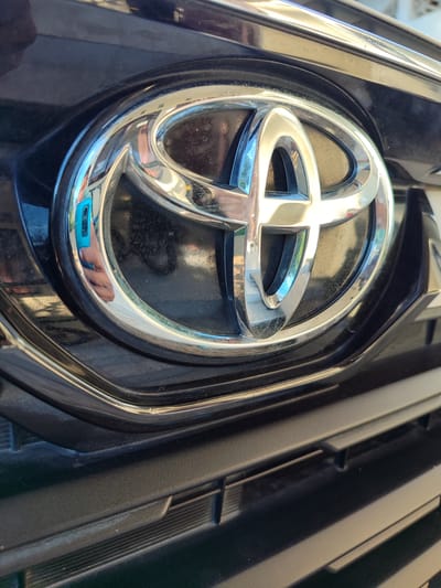 The image displays a close-up, low-angle shot of the Toyota emblem on the front grille of a vehicle. The emblem is silver and chrome, set against a black grille. The emblem is clean and reflects the surroundings, including what appears to be a blue phone and a hand. The background consists of the car's black body and grille. The image is taken outdoors, likely in sunlight, as indicated by the reflections and highlights. There is no discernible indication of the specific location within Lancokpanteara, Indonesia, beyond it being a place where a Toyota vehicle is present.
