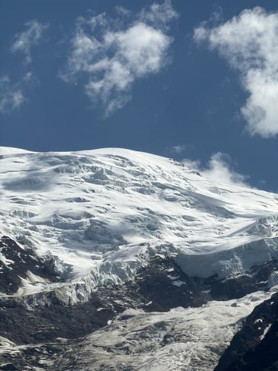 The image depicts a vast, snow-covered mountain range under a blue sky with scattered white clouds. The mountain's surface is textured with numerous crevasses and ridges, indicating a glacier. Dark, rocky terrain is visible at the base of the snow-covered slopes. In the upper left portion of the sky, a small, distant object, possibly a paraglider, is visible. The scene is likely a mountainous region, possibly the French Alps given the mention of Chamonix-Mont-Blanc, France, known for its glacial landscapes and mountaineering activities.