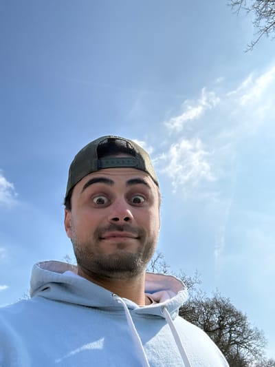 A male individual is centered in an upward-angled outdoor photograph, likely a selfie. He wears a light blue hooded sweatshirt with white drawstrings and an olive green mesh-back baseball cap positioned backwards. The man has dark hair and visible stubble/a short beard. His eyes are wide open, and his mouth is closed with a slight upward curve, creating an exaggerated or surprised expression directed at the camera. The background consists primarily of a clear blue sky with scattered, wispy white clouds, indicating bright daylight. In the lower right portion of the image, the bare branches of a deciduous tree are visible. The specific location within Kenilworth, United Kingdom, cannot be precisely identified from the visual data, but it is an outdoor natural environment.