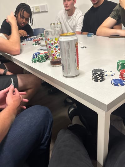 People are gathered around a white table, possibly playing a game like poker. Chips of various colors are scattered on the table. A can of "Belgium" beer and a decorated glass sit on the table. The participants include individuals wearing black and white shirts. One person has dreadlocks. A leg in blue jeans and another wearing athletic shorts are visible beneath the table. A pair of black and white shoes are underneath the table.