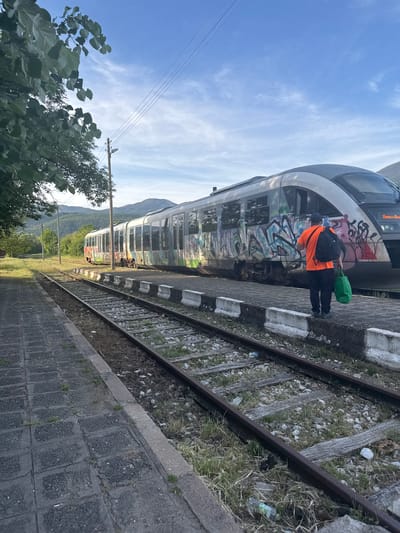 A train stands at a station in Bratsigovo, Bulgaria. The train is covered in graffiti, and a man in an orange shirt stands beside it with a black backpack and green bag. The scene is shot at ground level. The train is parallel to the tracks, and a platform edge is present on the right. Trash is visible around the railway tracks. Vegetation and distant mountains are visible. The sky is mostly clear with some clouds.