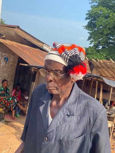 The scene is set in Amaseri, Nigeria, under a clear, bright blue sky with strong sunlight. The primary subject is an elderly Black man positioned in the foreground, facing left. He wears a dark blue long-sleeved shirt featuring textured horizontal stitching on the shoulders and a single circular gold-colored button at the collar opening. He has on round, light-tinted glasses with thin gold frames and a knitted hat with a white base, alternating black and orange horizontal stripes, and a prominent red, white, and black pom-pom on the left side of his head. His skin shows visible signs of aging.
In the mid-ground, to the left of the main subject, a Black woman is seated on a low structure or bench. She wears a patterned dress predominantly green with orange, black, and white designs. Behind her, two young Black children are seated; one wears a red top with white stripes, and the other wears a white top and light-colored bottoms, holding what appears to be a mobile phone. A third child is partially visible behind the woman. The woman wears pink sandals, and one of the children wears a green sandal, while another has bare feet. A faded picture or poster is affixed to the light-colored wall behind the seated individuals.
The background features a single-story building with a severely rusted corrugated iron roof and weathered walls, presenting an open doorway with a dark interior. A wooden beam extends horizontally above the doorway. To the far right, a makeshift structure composed of wooden poles and draped dark material is visible, with additional corrugated iron roofing. The ground is unpaved, consisting of dry, light-colored dirt. Green trees are present in the far background on the right.
