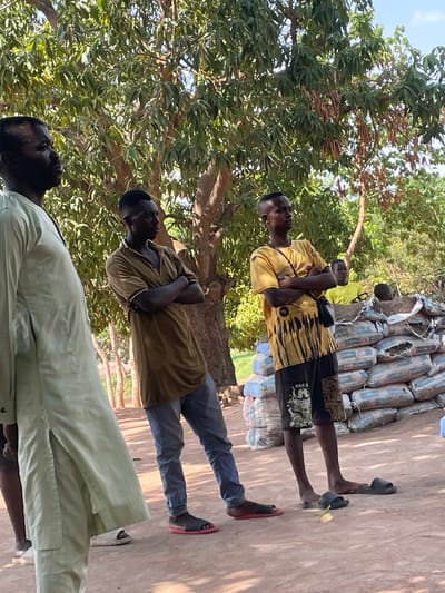 Three men stand outdoors, partially obscured by a tree. One man wears a light green shirt and pants, another wears a brown shirt and blue jeans, and the third wears a yellow patterned shirt and patterned shorts. There are stacked bags of cement behind one of the men. The location is Kado, Nigeria.