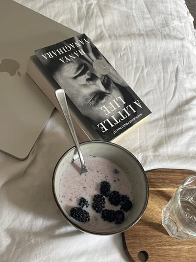 This media file presents a serene indoor scene, likely set on a bed covered with white, slightly crumpled linens. The arrangement suggests a moment of leisure, involving reading and a meal.
A prominent paperback book, titled "A Little Life" by Hanya Yanagihara, is featured. Its striking black and white cover displays a close-up of a man's contemplative face, with the text "MAN BOOKER PRIZE FINALIST" visible at the top. The book is positioned partly on a silver-grey laptop, of which the iconic Apple logo is partially visible at the bottom left.
Adjacent to the book is a rustic-looking ceramic bowl containing a light purple or pink liquid, resembling yogurt or a berry smoothie, generously topped with several plump, dark blackberries. A silver spoon rests within the bowl, indicating it's in use. To the upper right, an empty, clear glass with a textured, faceted design sits on a small, round wooden coaster.
The lighting is soft and ambient, suggesting a calm daytime atmosphere. The composition conveys a sense of quiet comfort and personal relaxation. This tranquil setting is situated in Aarhus, Denmark.