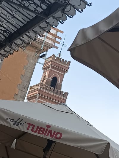 In Casalmaggiore, Italy, an outdoor cafe scene unfolds, dominated by a prominent bell tower. The tower, constructed of reddish-brown brick with white accents, features crenellated battlements and arched openings. A metal weather vane tops the structure. In the foreground, two large cafe umbrellas are partially visible. The left umbrella is white with the red text "caffè TUBINO" printed on it, along with a small pink heart symbol. The right umbrella is a muted beige color and is angled upward, obscuring a portion of the sky. A section of a building with a textured orange facade and stone accents is visible to the left of the tower. Above the building, a dark, ornate metal canopy with decorative scrollwork is present. The sky is a clear, pale blue. The image appears to be taken from a low angle, looking upwards towards the tower and buildings, with the umbrellas providing shade for cafe patrons. There are no people clearly visible in the frame.