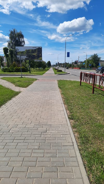 The image depicts a street scene in Smarhon, Belarus, on a sunny day with scattered clouds. The perspective is from a sidewalk paved with gray interlocking stones, which extends into the distance. To the left of the sidewalk is a strip of green grass, followed by a road with a few cars and streetlights. Beyond the road, trees and buildings line the street, suggesting a moderately developed urban or suburban area.

A prominent billboard is visible on the left side of the image, featuring a landscape photograph and text. The word "ЯКОР" (which translates to "Anchor" in English) is clearly legible on the billboard in large, dark letters. The billboard appears to be an advertisement or informational display.

In the background, more trees and structures are visible, along with a clear blue sky dotted with fluffy white clouds. The overall impression is one of a pleasant, ordinary day in a town, with elements of both nature and infrastructure present. There are no people visible in the image. The time of day is daytime, likely late morning or early afternoon, given the bright sunlight and shadows. The weather appears to be fair and warm.