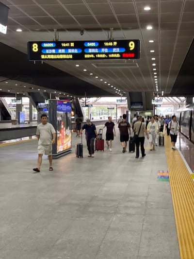 The image depicts a bustling train station platform in Longhua District, China. Several individuals, mostly dressed in casual attire, are waiting or walking along the platform. Some are seen with luggage, suggesting they are travelers. The scene appears to be indoors, under a covered structure, with artificial lighting from above. A yellow tactile paving runs along the edge of the platform, a common safety feature in public transportation areas.
A prominent digital display board shows train information. It indicates that train G6009, arriving at 16:41 from Changsha South, will terminate at Shenzhen North. Another train, G400, is scheduled to depart for Wuhan at 16:46 from Shenzhen North. The time of day is late afternoon, as indicated by the train schedules. The overall atmosphere seems calm but busy, with people engaged in their travel plans. The ground is paved with concrete tiles. The lighting suggests it could be daytime, but the covered nature of the station makes it difficult to ascertain the exact weather conditions. The presence of multiple train lines and the efficient display of information point to a well-developed public transport hub. The text on the signs is in Chinese, providing essential details for passengers.