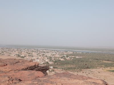 This panoramic image captures a sprawling landscape view of Dongola, Sudan, taken from a high vantage point, likely a rocky hill.
In the immediate foreground, reddish-brown rock formations dominate the lower left and bottom edges of the frame. These rocks exhibit rough, weathered textures and layered geological features, typical of arid, ancient terrain.
Moving towards the midground, a densely populated town or city unfolds, consisting of numerous light-colored buildings, predominantly low-rise structures, interspersed with scattered green trees. Roads and urban infrastructure are visible throughout the settlement. A few tall, slender communication towers can be seen rising above the buildings in the town area.
Beyond the town lies a wide, placid body of water, identifiable as the Nile River, which appears as a greenish-grey ribbon extending horizontally across the image. Its surface is calm, reflecting the hazy sky.
On the far bank of the Nile, an extensive and verdant expanse of date palm groves stretches across the landscape, indicating rich agricultural activity supported by the river. These groves form a dense, dark green band contrasting with the lighter tones of the town and distant arid land. Beyond the palm groves, the land transitions back to a lighter, arid terrain before fading into the distant, indistinct horizon.
The sky is a uniform, light blue-grey, appearing hazy, which suggests either atmospheric dust common to the region or a bright, diffused sunlight. The time of day appears to be daytime, possibly late morning or early afternoon, with no strong shadows. No specific human activity, individuals, or visible text can be discerned in the image. The scene portrays the symbiotic relationship between human settlement, agriculture, and the life-giving Nile River in an otherwise arid environment.