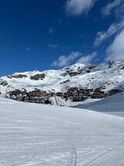 The image displays a panoramic view of the Val Thorens ski resort, located within the commune of Saint-Martin-de-Belleville, France. The foreground features a wide, snow-covered ski slope exhibiting parallel grooming patterns and individual ski tracks. In the midground, the village of Val Thorens is visible, comprising numerous multi-story buildings with modern alpine architecture, densely clustered along the lower mountain slopes. A snow-covered thoroughfare or ski run bisects the village, ascending into the terrain. The background is dominated by rugged, snow-capped mountain peaks and ridges, with exposed rock visible in areas. The sky is deep blue with scattered white cumulus clouds. The scene is brightly illuminated by direct sunlight. No individuals, vehicles, or specific actions are depicted.