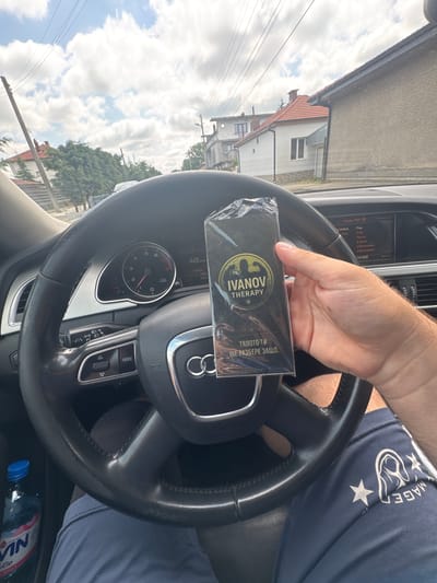 The image shows a first-person perspective from inside a car, looking towards the dashboard, steering wheel, and out the front windshield. A person's right hand is prominently displayed, holding a black, plastic-wrapped product package above the steering wheel.
**Subjects:** A person's hand and leg (wearing blue shorts), a house, trees, and cloudy sky.
**Objects:** An Audi steering wheel with the brand logo, a car dashboard with a speedometer (showing 0 km/h) and RPM gauge (at 0), an infotainment screen, a black packaged product, and a blue water bottle on the car floor.
**Activity or event taking place:** The person appears to be stopped or parked in a car, holding a product.
**Notable details:**
* **Time of day:** The infotainment screen shows "10:54", indicating late morning.
* **Weather:** The sky outside is bright and partly cloudy.
* **Location cues:** A typical house with a red-tiled roof and white walls is visible outside the car, alongside power lines and lush green trees, suggesting a residential or rural setting in Mogila, Bulgaria.
* **Emotions:** No specific emotions are discernible, the scene appears casual.
* **Car details:** The car is an Audi, identifiable by the steering wheel logo and dashboard design. The radio is set to "RONIKA" and displays categories like "Pop", "Folk", "News", "Events", and "Information".
**Visible Text:**
* On the product package: "IVANOV THERAPY" (with a golden logo) and in Bulgarian Cyrillic, "ТЯЛОТО ТИ ЩЕ РАЗБЕРЕ ЗАЩО." (which translates to "Your body will understand why.").
* On the infotainment screen: "Radio FM", "RONIKA", "Pop", "Folk", "News", "Events", "Information", "10:54", "Waveband", "Sound".
* On the person's shorts: Partially visible "IMAGE" and white stars.
* On the water bottle: Partially visible "NULA".