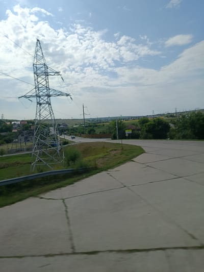 The image shows a bright, overcast day with a blue sky and scattered white clouds. A large, metallic electricity pylon dominates the left side of the frame. To the right, a paved road curves into the distance, bordered by a grassy embankment. In the background, a village with houses and trees is visible, nestled among rolling hills. Several other electricity pylons are scattered across the landscape, suggesting it is an area with power infrastructure. A black car is seen driving on the road in the mid-ground. The scene appears to be a rural or semi-rural area, possibly on the outskirts of a town.