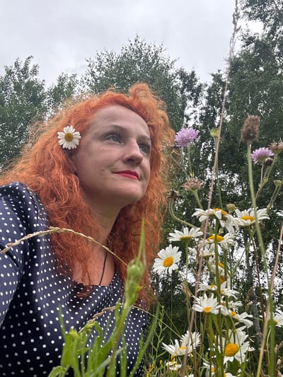 An outdoor, low-angle, medium shot captures a woman with bright orange, curly hair looking upwards and to the right. A white daisy with a yellow center is tucked behind her left ear. She wears a navy blue dress with white polka dots. Her head and shoulders are in focus against a backdrop of green trees and a cloudy sky. In the foreground and to the right, several daisies and purple wildflowers are in bloom. The location is the city of Privolzhsky, Russia.