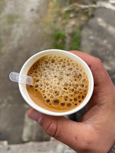 A person's right hand, with visible knuckles and tanned skin, holds a white disposable paper cup. The cup contains a dark brown liquid, identifiable as a beverage like coffee or espresso, covered by a thick, bubbly layer of amber-colored foam. A small, clear plastic stir stick is partially submerged in the liquid, resting against the inner rim of the cup. The background is heavily blurred, displaying indistinct tones of grey, brown, and green, consistent with an outdoor ground surface and minimal vegetation. The scene is located in Lehvaz, Armenia.