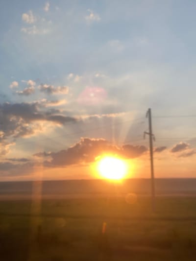 The image depicts a blurry sunset over a field in Balabanu, Moldova. The sun is low on the horizon, casting a warm orange glow across the sky and the land. The sky is filled with scattered clouds, some of which are illuminated by the setting sun. A utility pole stands to the right of the frame, with power lines stretching across the sky. The foreground is a blurred expanse of what appears to be a field. There are no people visible in the image. The overall impression is one of a tranquil, natural landscape at dusk.