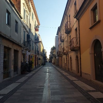 The image presents a ground-level view of a pedestrian street, identified as Corso Manthonè in Pescara, Italy, extending into the distance under a clear, light blue sky. The scene is illuminated by natural light, suggesting early morning or late afternoon, with sunlight primarily hitting the upper sections of the buildings and casting long shadows across the pavement.

On the left, multi-story buildings, predominantly light grey and beige, feature various windows, some with closed green or light-colored shutters, and several balconies with dark metal railings. The ground floor exhibits commercial facades, some with security roller shutters lowered, and potted plants are visible near entrances.

On the right, buildings appear in shades of orange and light yellow, also multi-story, with multiple windows and balconies featuring dark metal railings. Arched entrances are present on the ground level, some fitted with dark double doors; a faded building number is discernible near one such entrance.

The street itself is paved with an intricate pattern of light grey and darker patterned flagstones, creating a distinct central pathway and bordered sections. A prominent, lighter-colored strip runs down the center of the main pedestrian area. The street appears largely devoid of human activity; a single, highly indistinct figure is faintly visible in the far distance. Overhead, a thin, dotted line stretches horizontally across the street between the buildings.