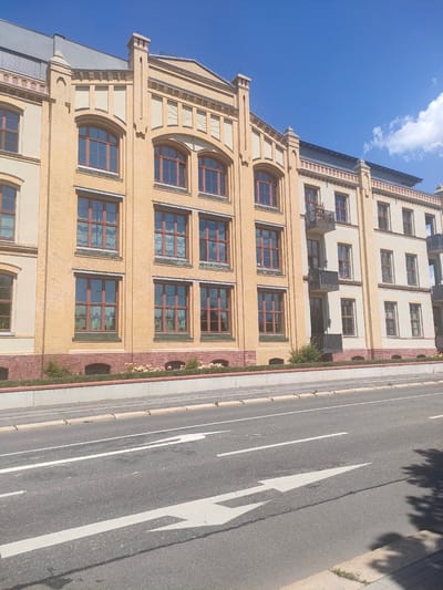 The image depicts a building in Chemnitz, Germany, with a street in the foreground. The building is light yellow brick with decorative red brick accents and numerous windows. It appears to be an apartment building or commercial space with multiple floors. There are balconies on some of the upper floors. A grey road with white lane markings and an arrow indicating direction runs in front of the building. The sky is blue with a few white clouds. The scene is captured in daylight.