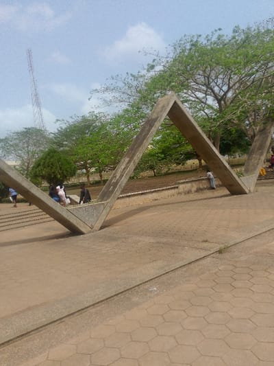 The image captures an outdoor scene in Adzho, Nigeria, under a partly cloudy blue sky. The foreground features an expanse of paved ground constructed from hexagonal concrete pavers, bisected by a narrow, raised concrete curb. Dominating the central midground is a large, weathered concrete sculpture composed of interconnected angled beams, forming a series of geometric "V" or chevron shapes that extend into and out of the ground. In the midground, several individuals are present: two figures walk on a paved path to the left of the sculpture; a small group of three people, including one in a black top and another in a light-colored dress, are gathered under the left arch of the concrete structure; and a single person in a light shirt leans against a low concrete wall under the right arch. The background consists of a treeline with dense green foliage, indicating a park-like or campus environment. A tall, lattice-style communications tower is visible in the far left background, rising above the trees. Shadows are cast by the concrete sculpture and individuals, consistent with daylight illumination.