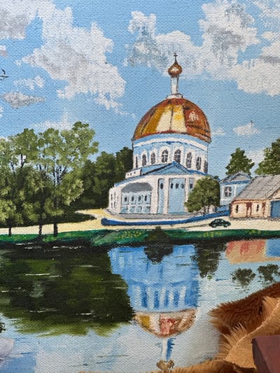The painting depicts a view of the Church of the Ascension in Rasskazovo, Russia. The church has a prominent golden dome topped with a cross, and a white facade with blue accents. It is surrounded by lush green trees and a body of water in the foreground. The water reflects the church and the sky, which is filled with fluffy white clouds against a bright blue backdrop. There are also smaller buildings visible to the right of the church, and a dark-colored car is parked on a path near the church. The overall scene is idyllic and peaceful, capturing the beauty of the church and its natural surroundings.