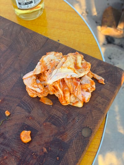 A pile of seasoned chicken skins sits on a wooden cutting board, which is on a yellow table. The chicken skins appear to be raw and coated in a reddish-orange spice mixture and black pepper. A small piece of something orange, possibly a piece of carrot, is to the left of the pile of chicken skins. In the upper left corner, part of a glass bottle with a yellow liquid and a label with Cyrillic text is visible. The background is out of focus, showing a sandy area and what appears to be legs and feet. The scene is likely outdoors, possibly in a park or backyard setting. The location is specified as Povarovka, Russia.