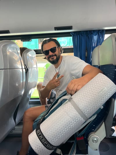 An individual with dark hair and a beard, wearing sunglasses and a white t-shirt, sits in a bus seat. They are holding up two fingers in a peace sign and have a rolled-up sleeping mat, labeled "GTXSPORT", strapped to their backpack. The interior of the bus is visible, with blue curtains and gray seats. Outside the window, a green tarp and building are visible, suggesting an urban environment, possibly La Llagosta, Spain.