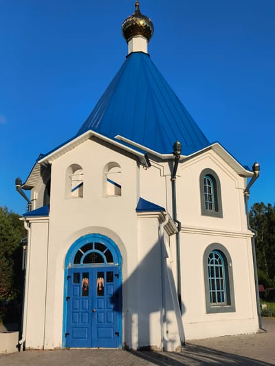 A bright white, small Orthodox chapel with a blue, steeply pitched roof and a golden dome stands under a clear blue sky in Aya, Russia. The chapel has a prominent, arched blue double door at the front, with two panels on each door displaying stylized images of winged figures, possibly angels, in red and gold. Above the doors is a semicircular window with a cross design. To the sides of the main entrance, there are arched windows. The building features decorative gutters with golden finials. The chapel is situated on a paved area, and trees are visible in the background. The lighting suggests it is daytime, with shadows cast on the building and ground.