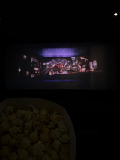 A movie screen illuminates the dark theater with an abstract cityscape, blurring details. To the right, a partially visible bucket overflows with popcorn, bathed in the faint glow. A single light source dots the upper left corner, further accentuating the darkened environment. The scene captures the quintessential movie-going experience.