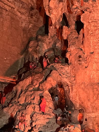 The image depicts a scene within a cave in Gollagutta, India. The cave walls are rough and textured, illuminated by an orange light that casts deep shadows. Several figures, appearing to be statues or mannequins, are positioned throughout the cave.
In the foreground, a seated figure with light skin and dark hair is dressed in red garments. To the right and slightly behind him, another figure in red is standing on a rocky outcrop, facing away from the viewer. Above them, a figure in a patterned garment is seen ascending a rocky path.
Further into the cave, there are more figures. One figure in red is positioned on a ledge to the left. In the background, within a darker recess of the cave, several figures are clustered together, some appearing to be seated. One figure in a blue shirt is standing in this recess, looking into the depths of the cave.
The environment is natural and cavernous, with stalactite-like formations visible on the cave ceiling and walls. The overall impression is of a staged scene within a natural setting, possibly a religious or historical exhibit.