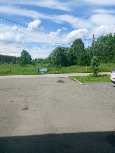This image depicts an outdoor scene on a sunny day, likely in Volchansk, Russia, characterized by a clear blue sky with scattered white clouds and a line of trees in the background. In the foreground, there is a paved area, possibly a parking lot or road, with some dark patches indicating stains or wear. A strip of green grass borders the paved area, and further back, a lush green field stretches towards the trees.
Visible elements include:
**Objects:**
* A metallic structure, possibly a bench or a small shelter, is situated in the grassy area.
* A wooden utility pole stands to the right of the frame, with wires extending from it.
* A white vehicle is partially visible on the right side of the image, showing its rear bumper and a license plate.
* A yellow marker or sign is present in the grassy area near the utility pole.
**Environment:**
* The sky is bright blue with cumulus clouds, suggesting fair weather.
* The foreground is dominated by an asphalt surface.
* The midground features a field of green grass, with a line of dense trees forming the backdrop.
* The overall impression is one of a rural or semi-rural setting, with elements of human infrastructure integrated into the natural landscape.
There are no people visible in the image. The scene is static, with no apparent actions or interactions taking place. The lighting suggests it is daytime, with the sun casting shadows that are partially visible in the lower part of the frame.