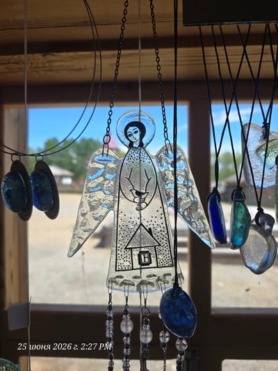 The image depicts an outdoor scene with a focus on several glass ornaments hanging from a wooden structure. The primary subject is a translucent glass angel with black painted details, including a halo, facial features, wings, and a depiction of a house on its body. The angel is suspended by a dark chain. To its left, two circular glass pendants with dark metal frames hang. To the right of the angel, various other glass pendants of different shapes and colors, including blue, green, and clear glass, are visible, suspended by black cords. In the background, a blurred view of a sunny outdoor environment is visible, featuring greenery, a building with a red roof, and a bright blue sky. A timestamp in Russian, "25 июня 2026 г. 2:27 PM", is overlaid on the lower left of the image. The overall impression is of a souvenir shop or a display area in a cultural or historical site, possibly related to the location "Тальцы (иркутский), Russia," which is a well-known open-air museum of architecture and ethnography.