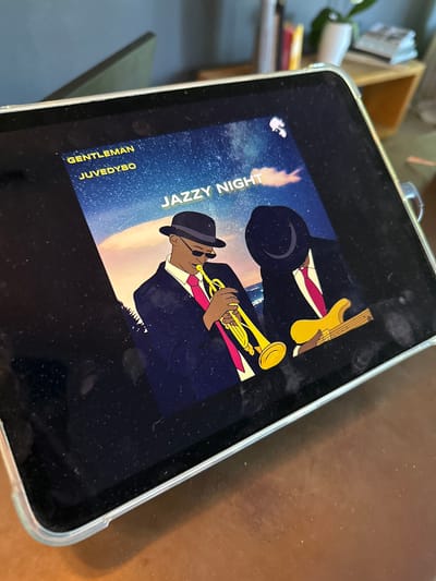 A tablet computer displays album art featuring two cartoon figures in suits and hats. The figure on the left plays a trumpet, while the figure on the right holds a yellow electric guitar. The background depicts a starry night sky with clouds and a sunset over water. Text on the album art reads "GENTLEMAN," "JUVEDY.BO," and "JAZZY NIGHT." The tablet is placed on a wooden surface, and the background shows a blurred room interior with shelves and a plant. The image does not provide any information about the city of St. Märgen, Germany.