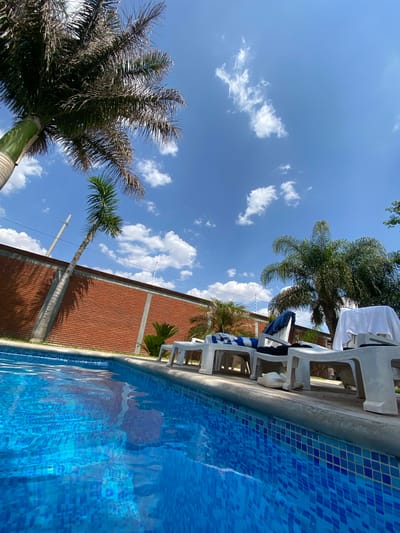 The image depicts a swimming pool area in San Francisco Lachigoló, Mexico. The scene includes a partially visible blue-tiled swimming pool, with a concrete edge. Several white plastic lounge chairs are positioned along the pool's edge, some with towels draped over them. A brick wall runs parallel to the pool. Palm trees are visible in the background, against a clear blue sky dotted with fluffy white clouds. No people are visible in the frame.